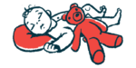 An infant sleeps with their head on a red pillow while a red teddy bear rests on their stomach.